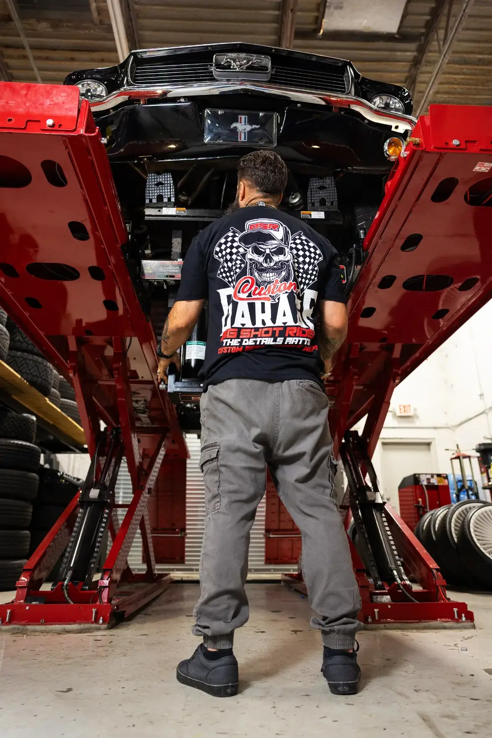 Chris Joerger under a classic Mustang on the lift at Big Shot Rides &mdash; shirt reads The Details Matter